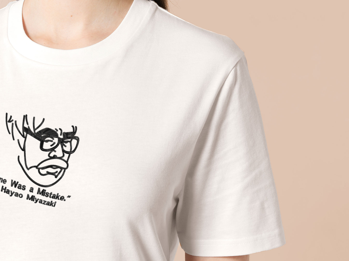 A off white crew neck short sleeve t-shirt, with an embroidered black thread design of a portrait of a depressed Hayao Miyazaki the creator of Studio Ghibli with text underneath that reads "Anime Was A Mistake." - Hayao Miyazaki