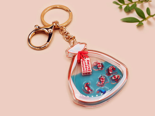 A large transparent acrylic shaker keychain with gold keyring, filled with mini chibi shrimps and labelled as emotional support shrimp