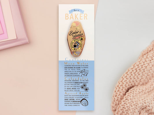 Gold Enamel Talisman Pin with yellow design and the words Baker's Talisman sits on a long white and blue backing card with gold accents. The backing card has details the symbolism of the different design elements of the Talisman pin.