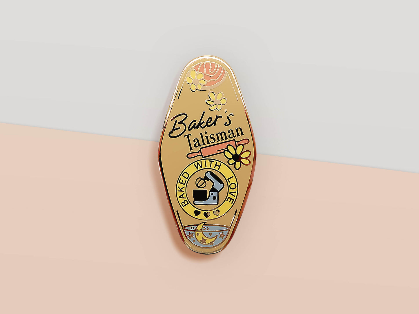 Gold Enamel Talisman Pin with yellow design and the words baker's Talisman, Baked With Love. The pins design includes a mixer and a rolling pin, moon water, as well as flowers and crystals.