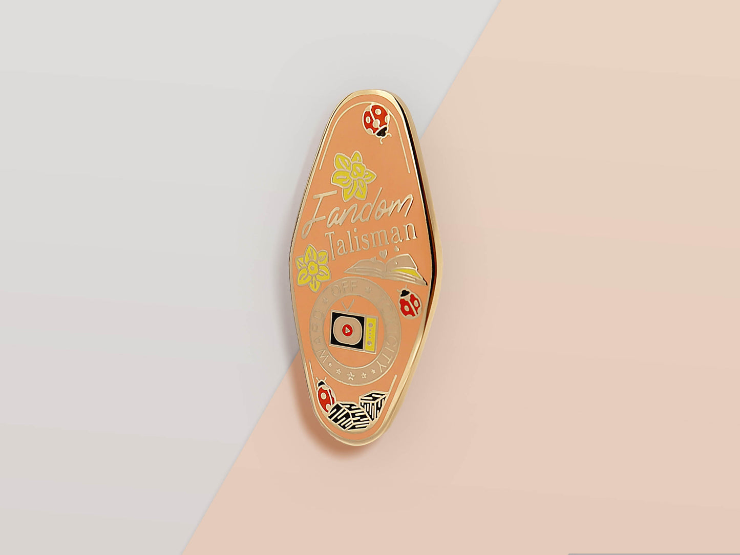 Gold Enamel Talisman Pin with pink design and the words Fandom Talisman, Ward of toxicity block. The pins design includes a television and an open book, ladybugs, as well as flowers and crystals.