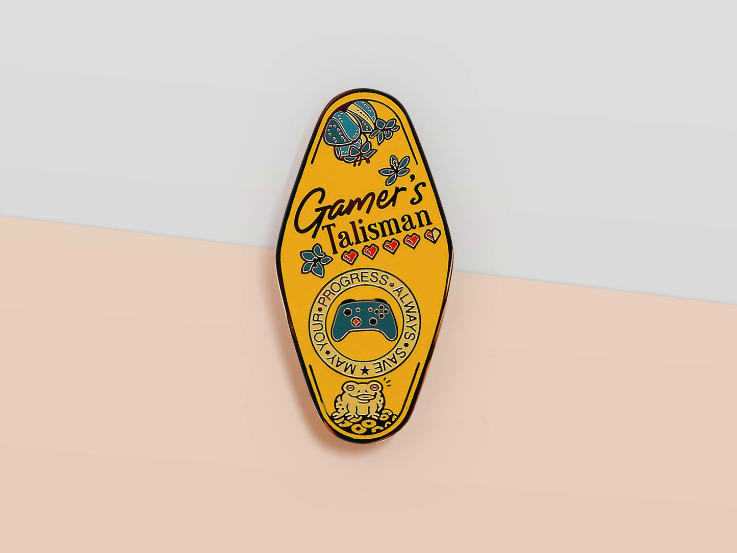 Gold Enamel Talisman Pin with yellow design and the words Gamer's Talisman, May Your Progress Always Save. The pins design includes a game console controller and health hearts, A jin Chan money toad, as well as plants and crystals.