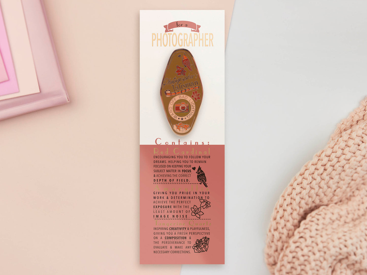 Gold Enamel Talisman Pin with brown design and the words Photographer's Talisman sits on a long white and red backing card with gold accents. The backing card has details the symbolism of the different design elements of the Talisman pin.