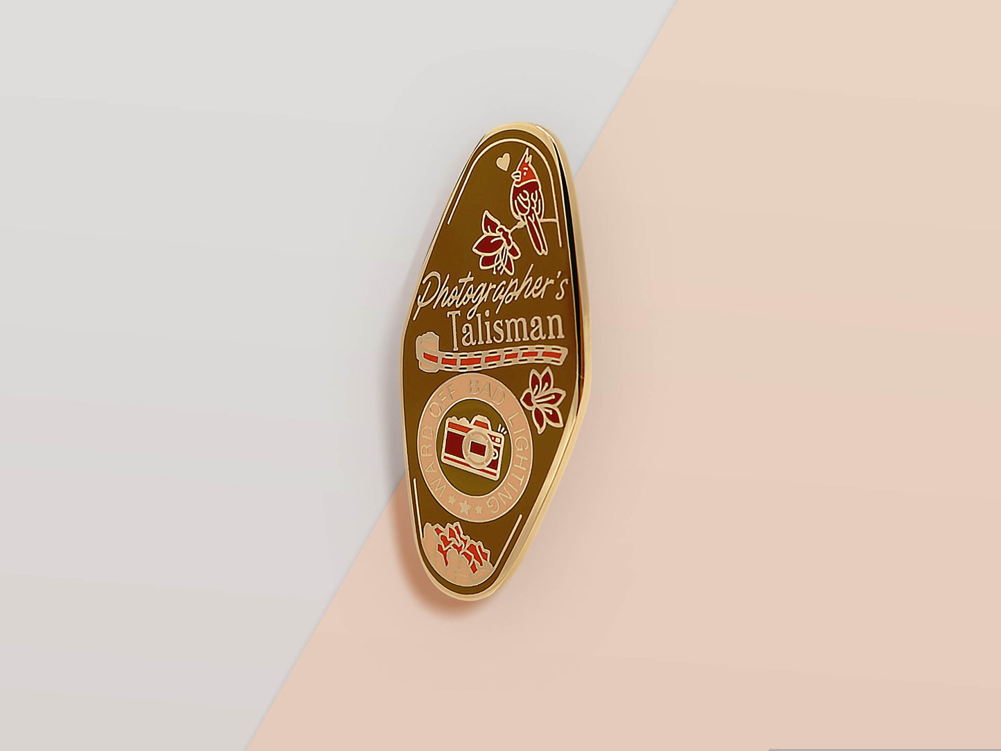 Gold Enamel Talisman Pin with brown design and the words Photographer's Talisman, Ward of bad lighting. The pins design includes a camera and a film, a red bird, as well as flowers and crystals.