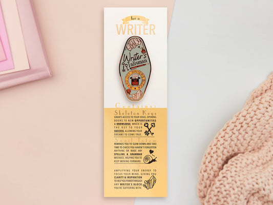 Gold Enamel Talisman Pin with blue design and the words Writer's Talisman sits on a long white and yellow backing card with gold accents. The backing card has details the symbolism of the different design elements of the Talisman pin.