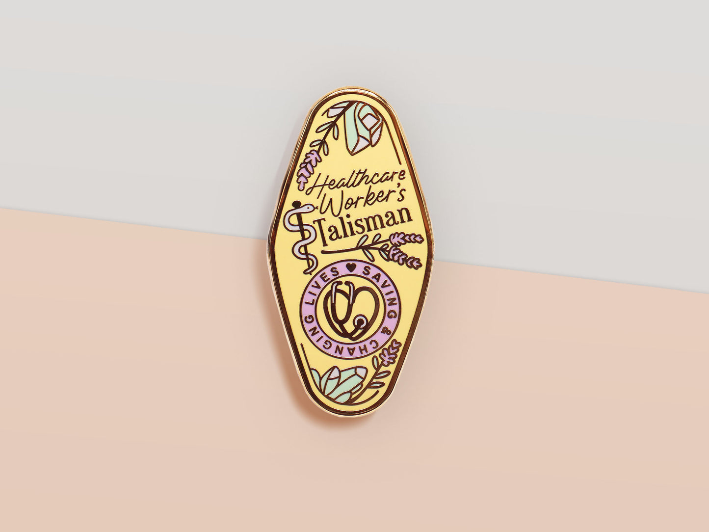 A Health Worker's Talisman gold enamel pin on a gold foiled backing card that outlines the symbolism of the talisman including information and imagery on the Rod of Asclepius, Lavender Stems and Prehnite Crystals