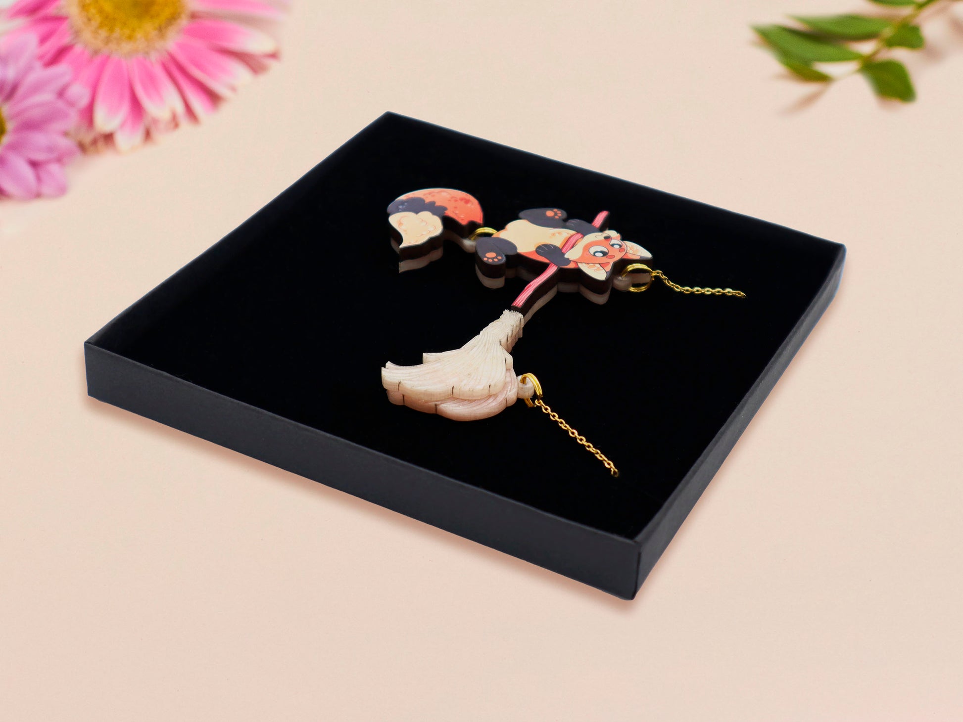 Mixed material handmade necklace of chibi cartoon Fox hanging on to a pearlescent witches broomstick, with a gold chain and black gift box.