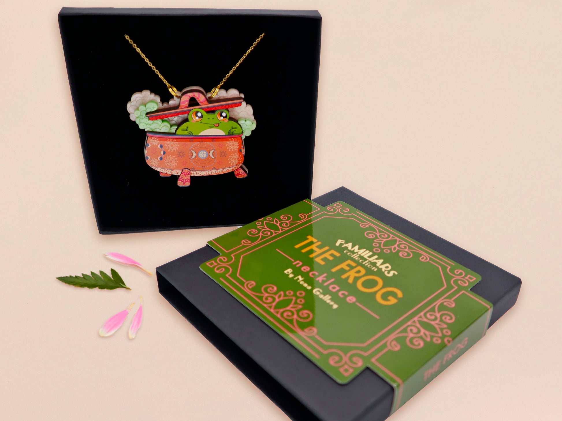 Mixed material handmade necklace of chibi cartoon frog sat inside a bubbling pearlescent witch's cauldron, with a gold chain and black gift box with a green familiars collection gift sleeve.