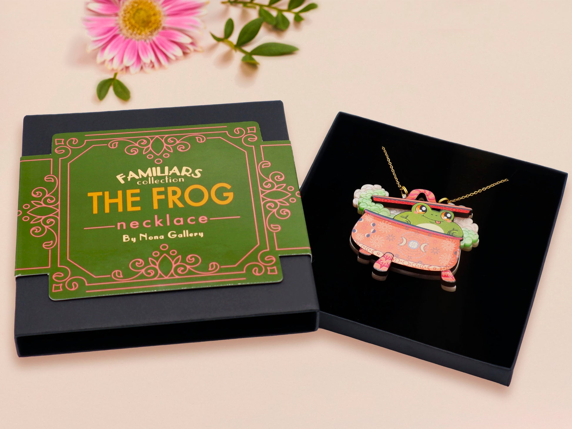 Mixed material handmade necklace of chibi cartoon frog sat inside a bubbling pearlescent witch's cauldron, with a gold chain and black gift box with a green familiars collection gift sleeve.