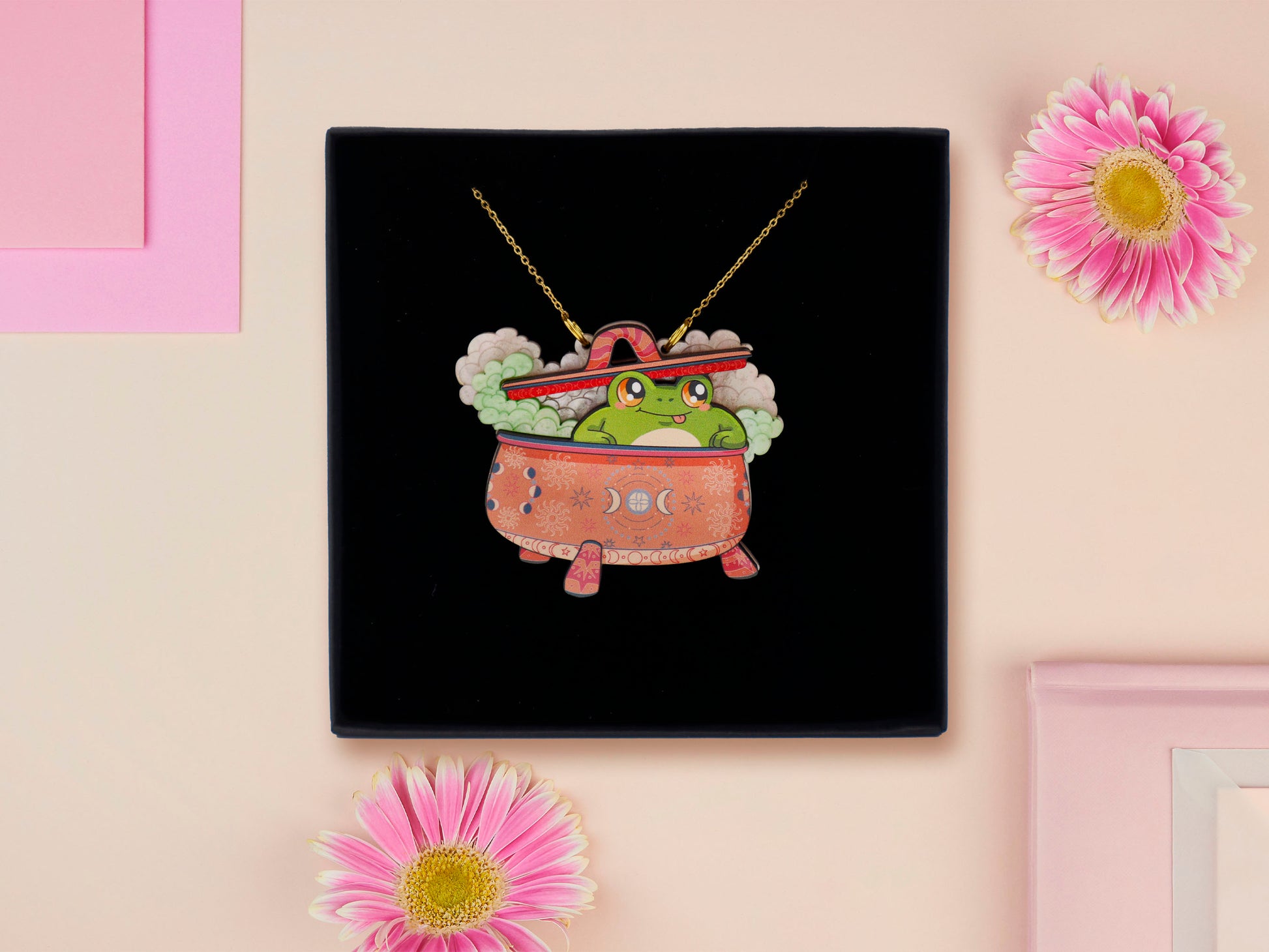 Mixed material handmade necklace of chibi cartoon frog sat inside a bubbling pearlescent witch's cauldron, with a gold chain and black gift box.