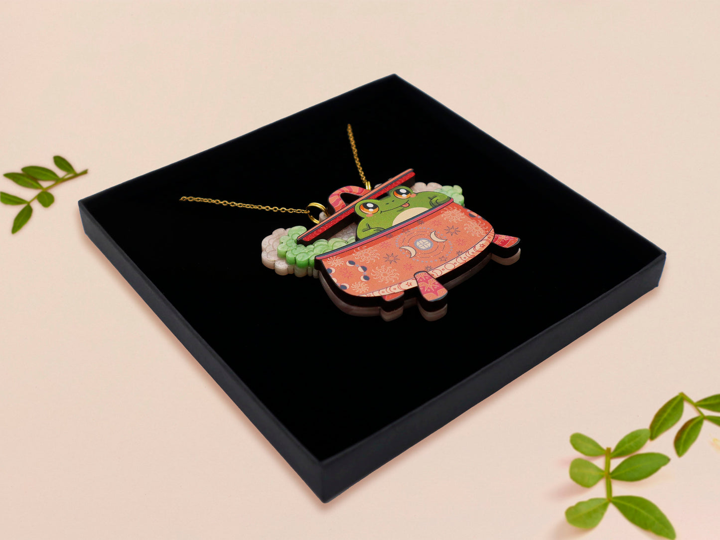 Mixed material handmade necklace of chibi cartoon frog sat inside a bubbling pearlescent witch's cauldron, with a gold chain and black gift box.