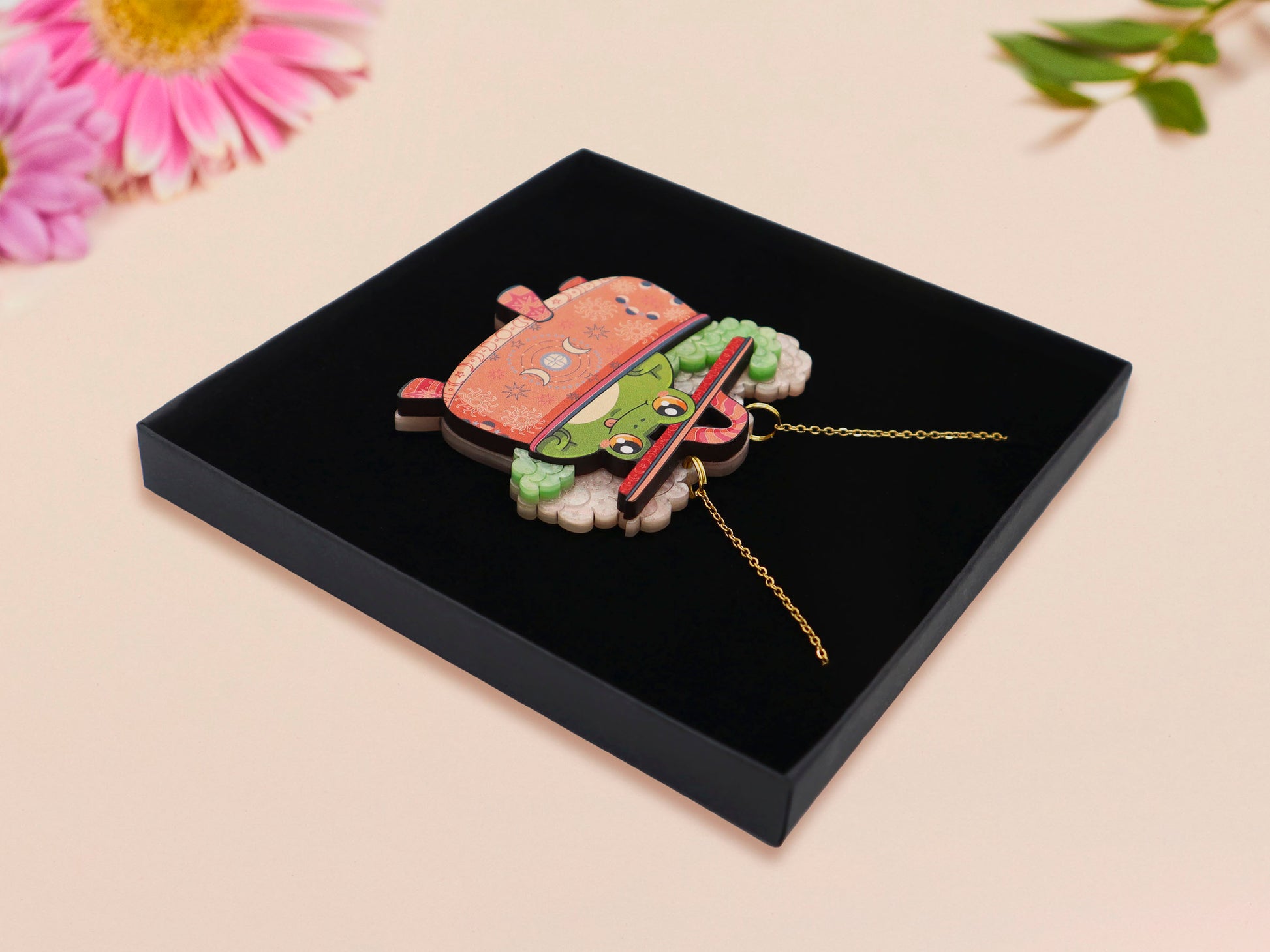 Mixed material handmade necklace of chibi cartoon frog sat inside a bubbling pearlescent witch's cauldron, with a gold chain and black gift box.