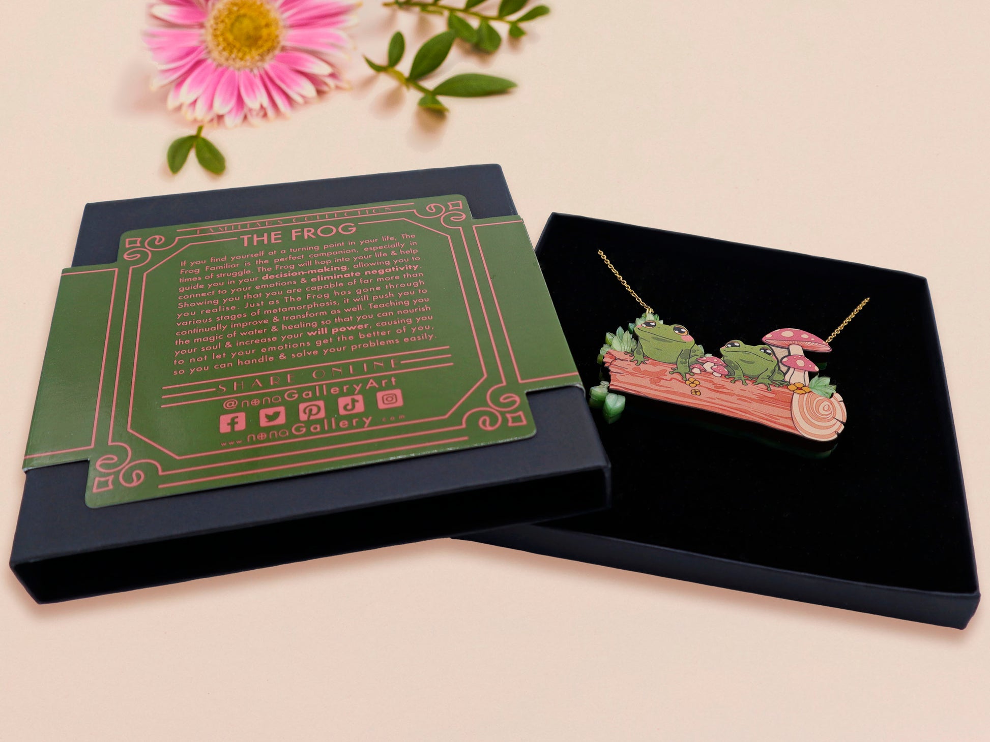 A wooden and glitter acrylic necklace with gold chain in a black box of two cute frogs sat on a log smiling surrounded by flowers, plants and mushrooms