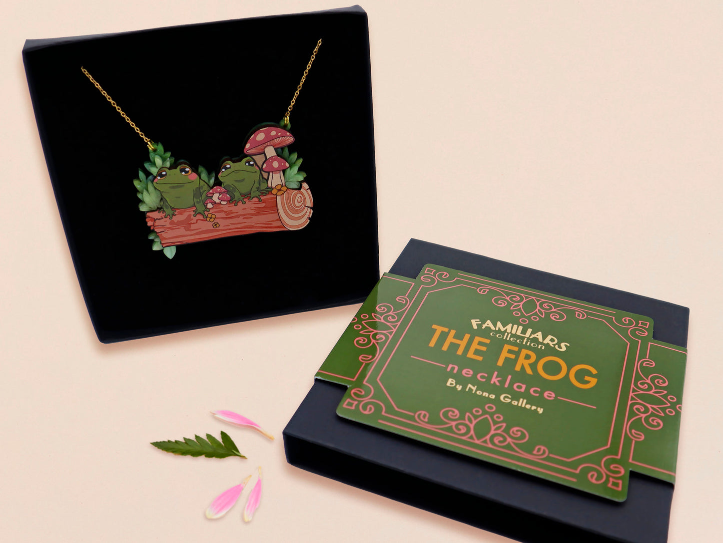 A wooden and glitter acrylic necklace with gold chain in a black box of two cute frogs sat on a log smiling surrounded by flowers, plants and mushrooms