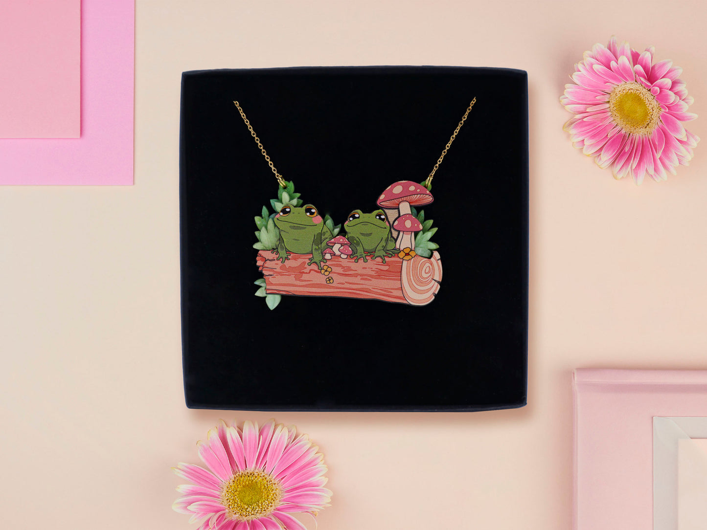 A wooden and glitter acrylic necklace with gold chain in a black box of two cute frogs sat on a log smiling surrounded by flowers, plants and mushrooms