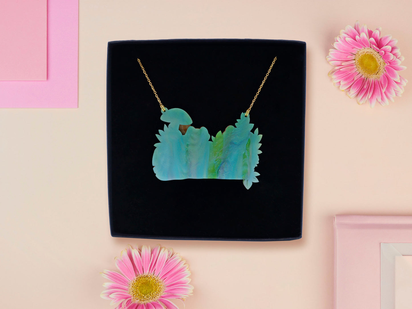 A wooden and glitter acrylic necklace with gold chain in a black box of two cute frogs sat on a log smiling surrounded by flowers, plants and mushrooms