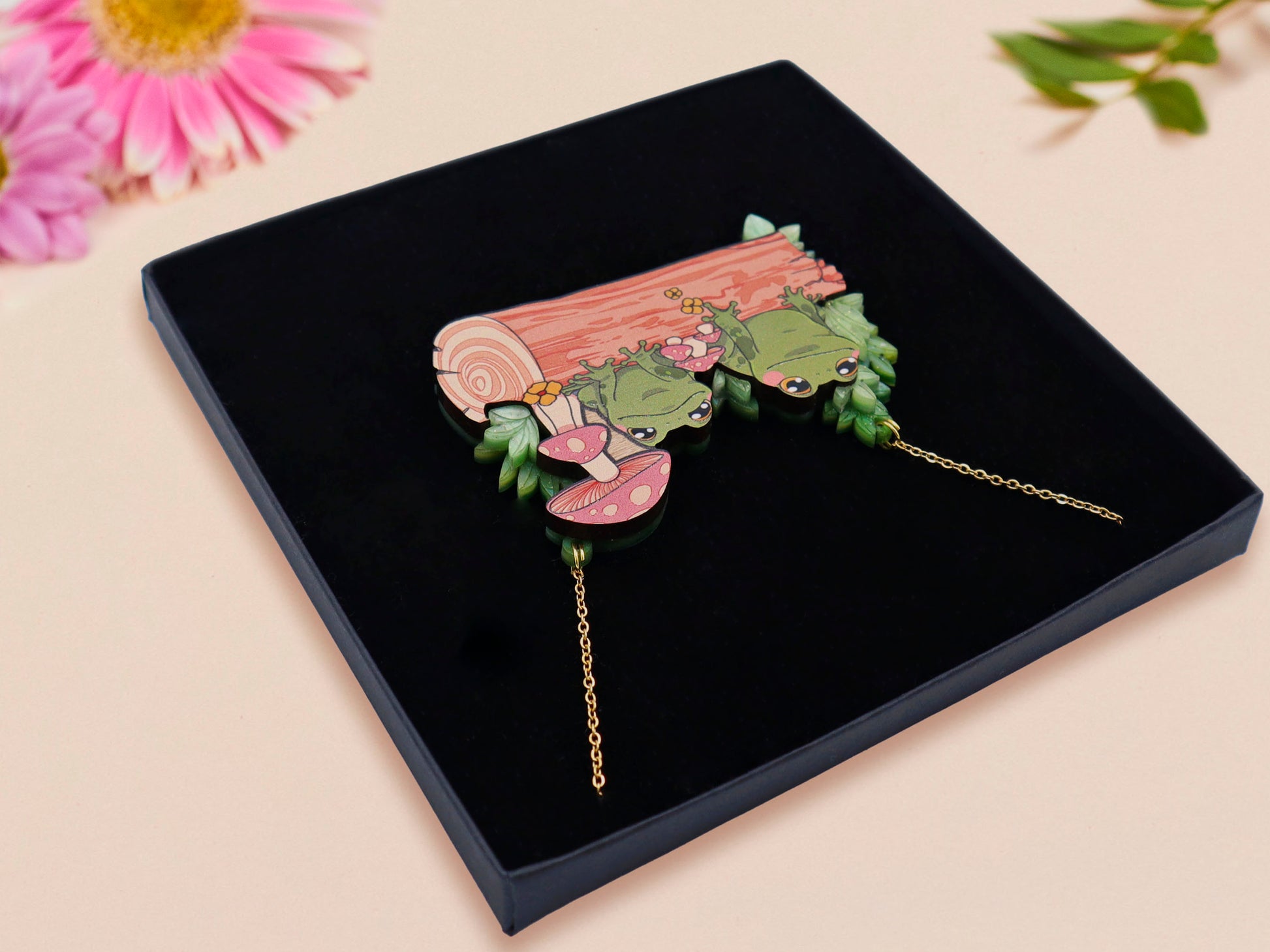 A wooden and glitter acrylic necklace with gold chain in a black box of two cute frogs sat on a log smiling surrounded by flowers, plants and mushrooms