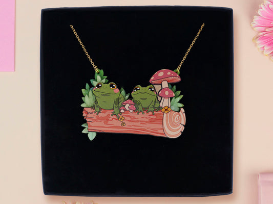 A wooden and glitter acrylic necklace with gold chain in a black box of two cute frogs sat on a log smiling surrounded by flowers, plants and mushrooms