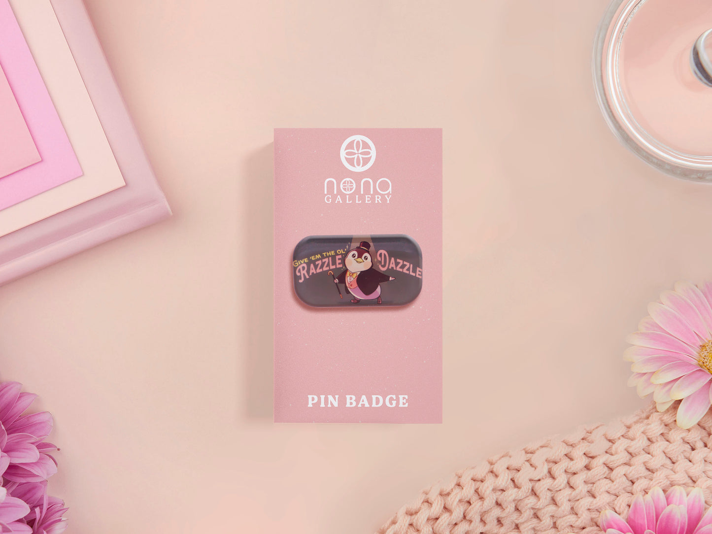 A clear acrylic pin badge with a pink heart-shaped pin back clasp. The design of the pin badge is of a cute chibi penguin dancing in a suit under a spotlight with the quote give 'em the ol' razzle dazzle