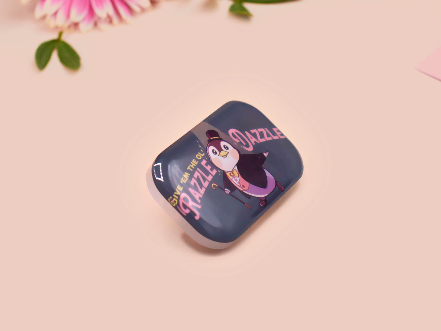 A clear acrylic pin badge with a pink heart-shaped pin back clasp. The design of the pin badge is of a cute chibi penguin dancing in a suit under a spotlight with the quote give 'em the ol' razzle dazzle