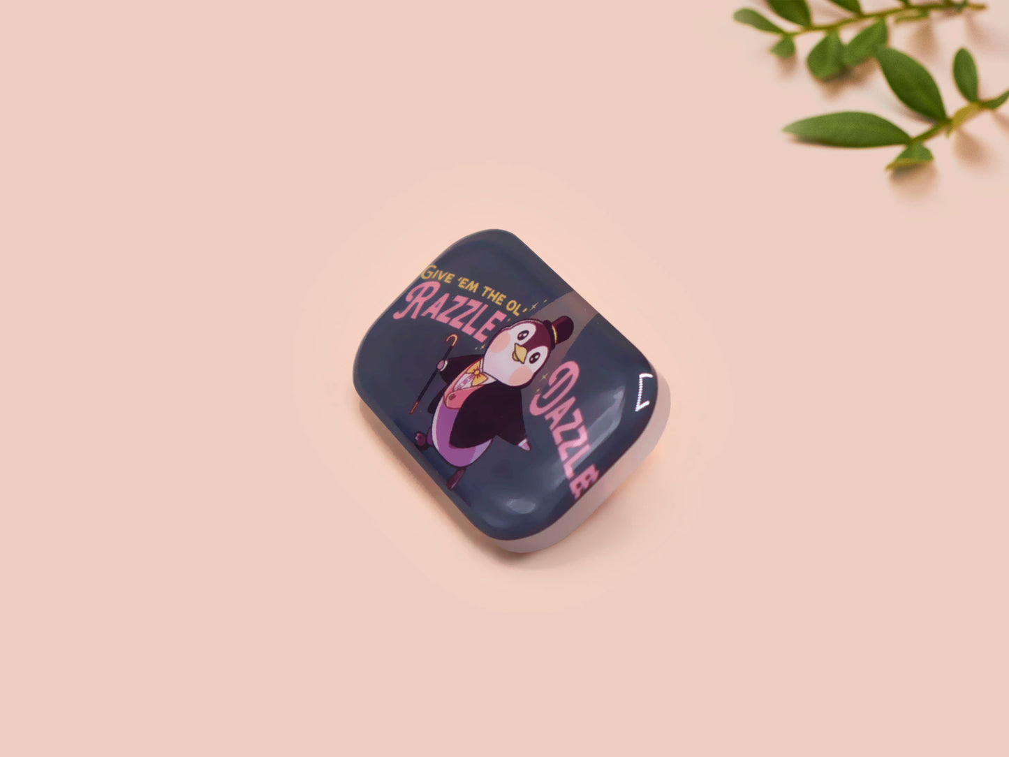 A clear acrylic pin badge with a pink heart-shaped pin back clasp. The design of the pin badge is of a cute chibi penguin dancing in a suit under a spotlight with the quote give 'em the ol' razzle dazzle