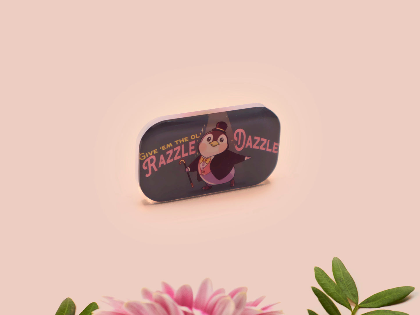 A clear acrylic pin badge with a pink heart-shaped pin back clasp. The design of the pin badge is of a cute chibi penguin dancing in a suit under a spotlight with the quote give 'em the ol' razzle dazzle