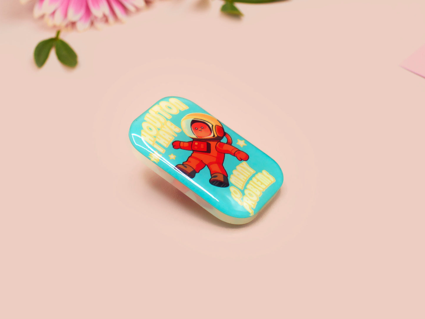 A clear acrylic pin badge with a pink heart-shaped pin back clasp. The design of the pin badge is of a cute chibi retro astronaut floating in sapce with the quote Houston I have so many problems