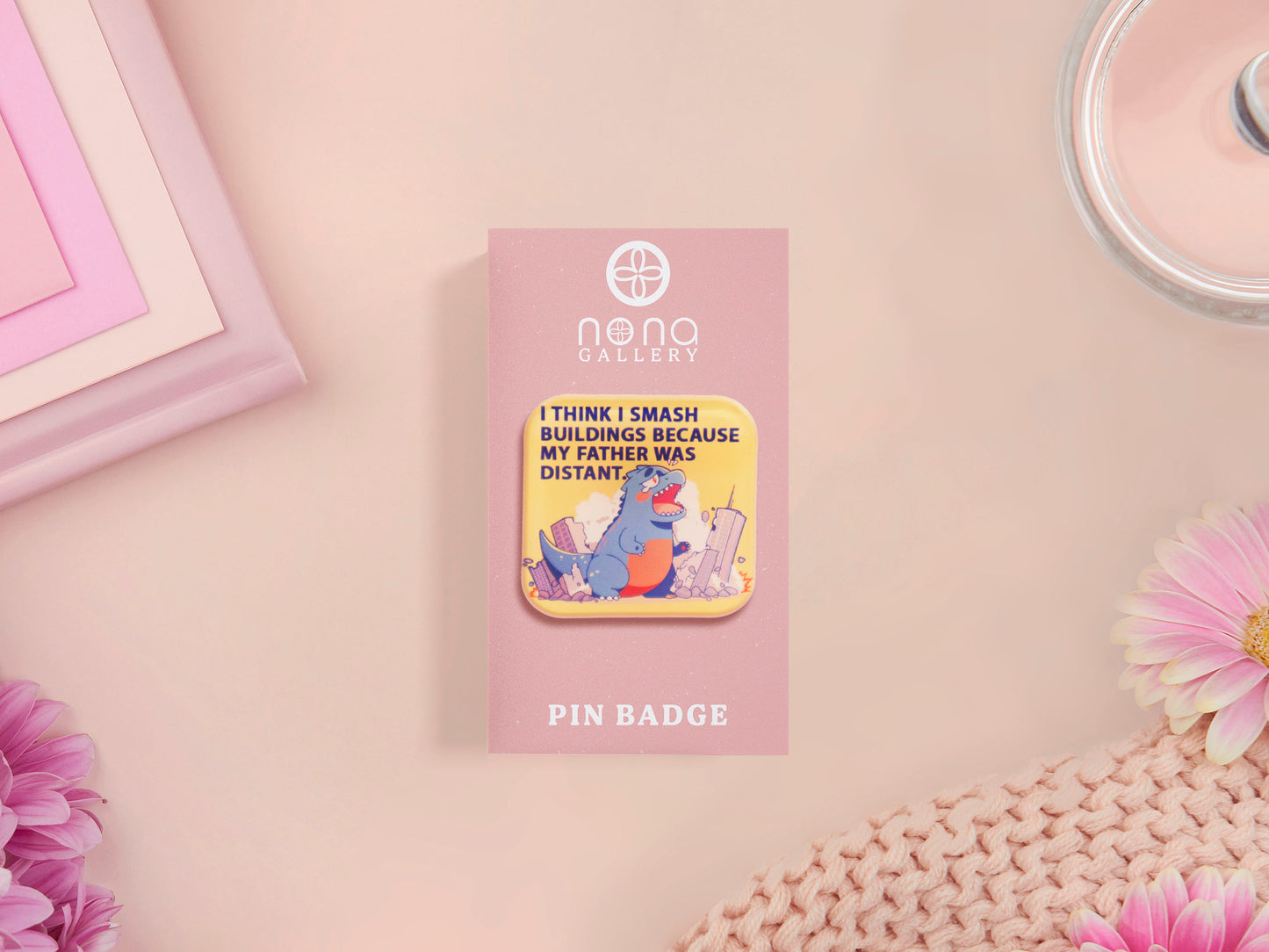 A clear acrylic pin badge with a pink heart-shaped pin back clasp. The design of the pin badge is of a cute chibi blue godzilla like kaiju destroying a city with the quote I Think I Smash Buildings Because My Father Was Distant.
