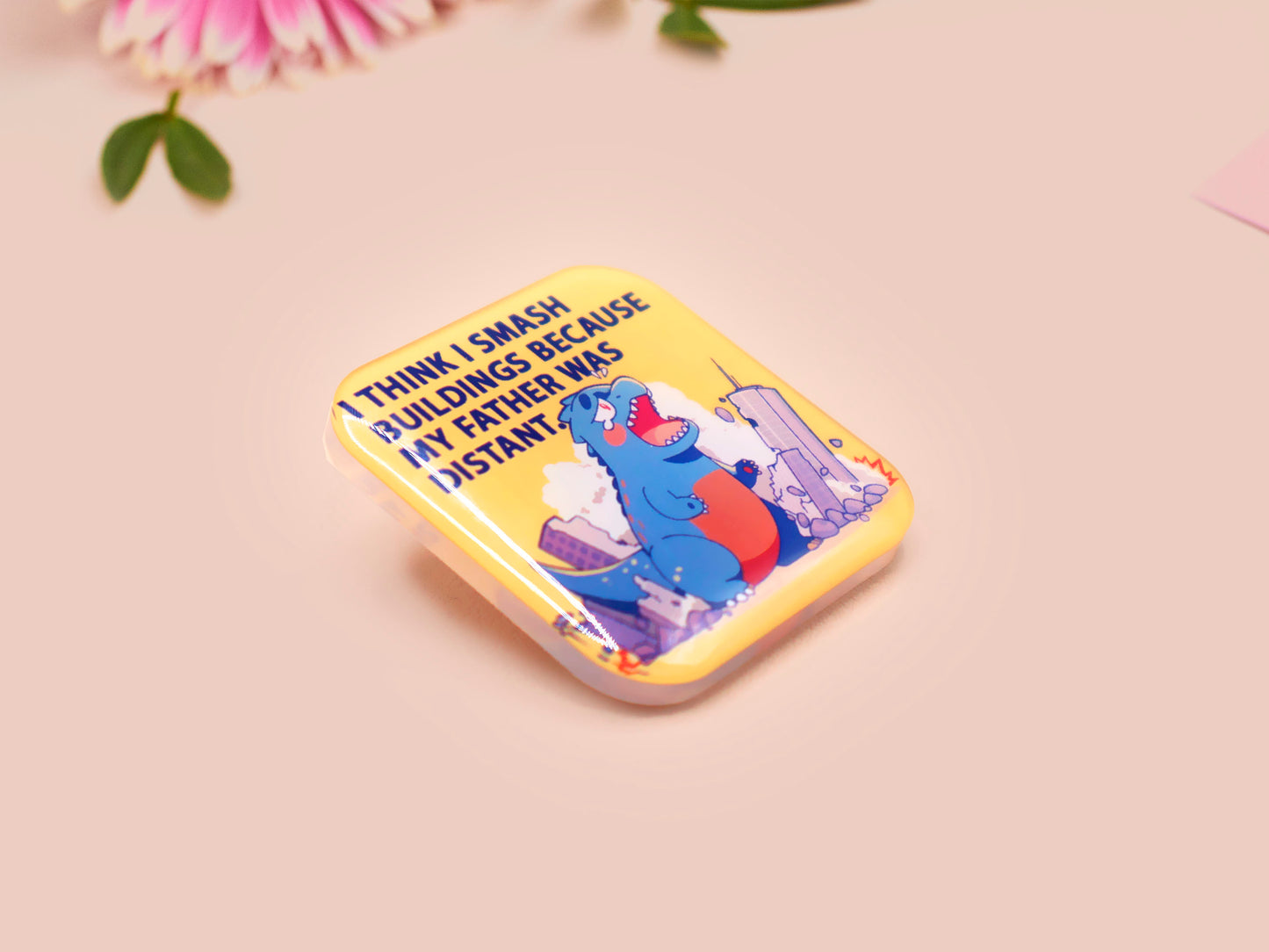A clear acrylic pin badge with a pink heart-shaped pin back clasp. The design of the pin badge is of a cute chibi blue godzilla like kaiju destroying a city with the quote I Think I Smash Buildings Because My Father Was Distant.