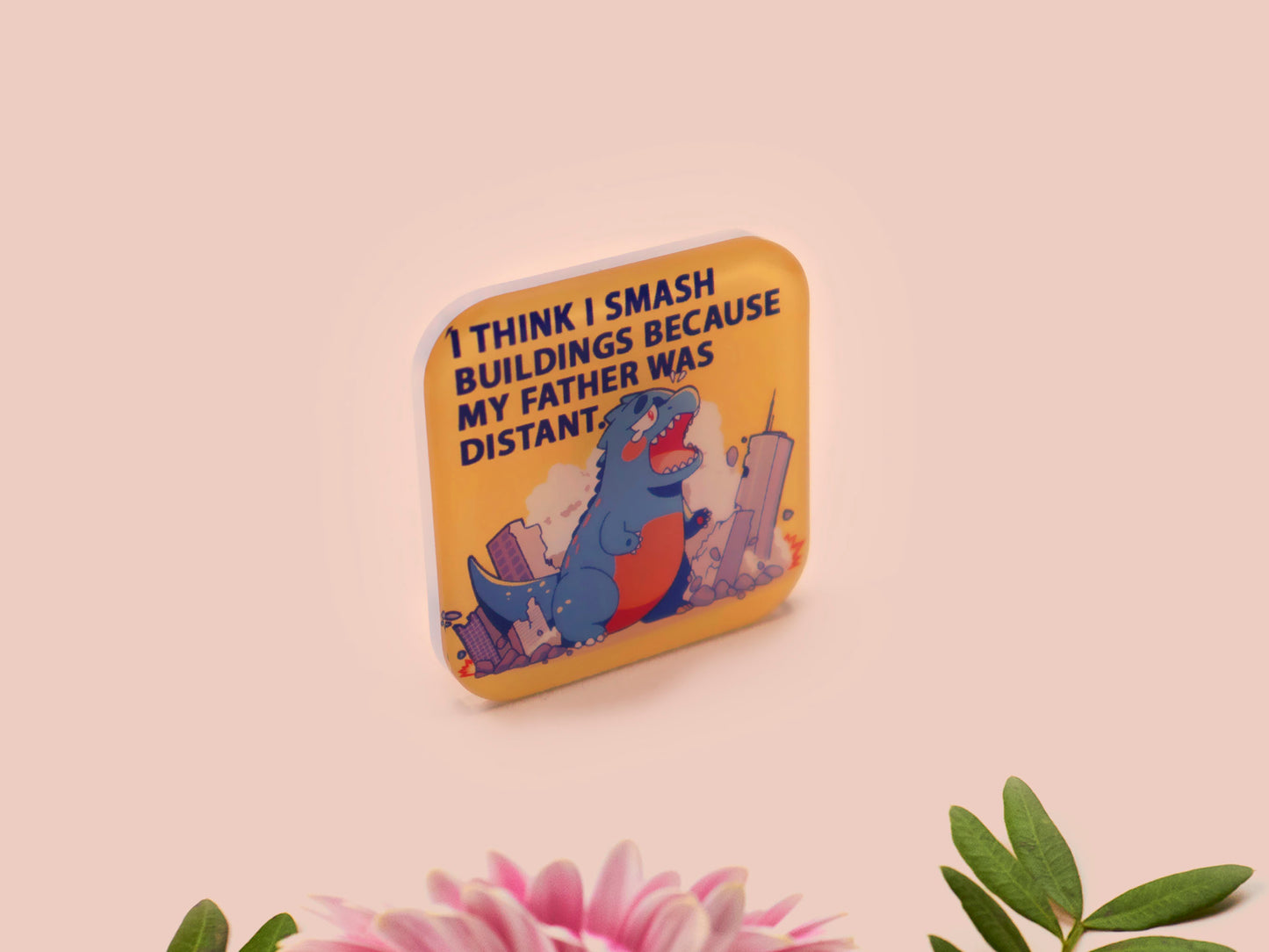 A clear acrylic pin badge with a pink heart-shaped pin back clasp. The design of the pin badge is of a cute chibi blue godzilla like kaiju destroying a city with the quote I Think I Smash Buildings Because My Father Was Distant.