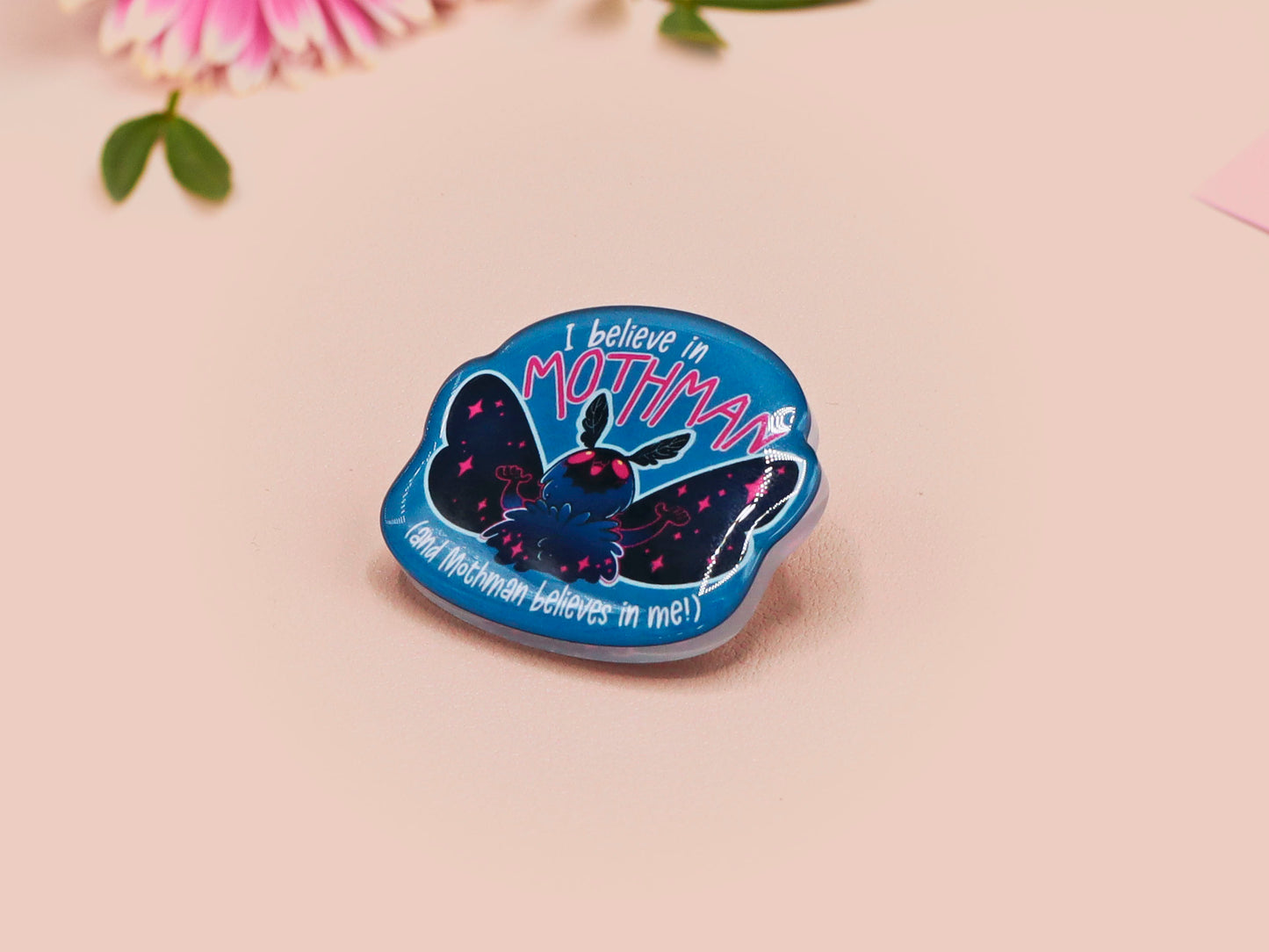 Clear acrylic pin badge with pink heart-shaped rubber pin back clasp. The design of the pin badge is a cute cartoon chibi Mothman cryptid with the quote I believe in Mothman and Mothman believes in me.