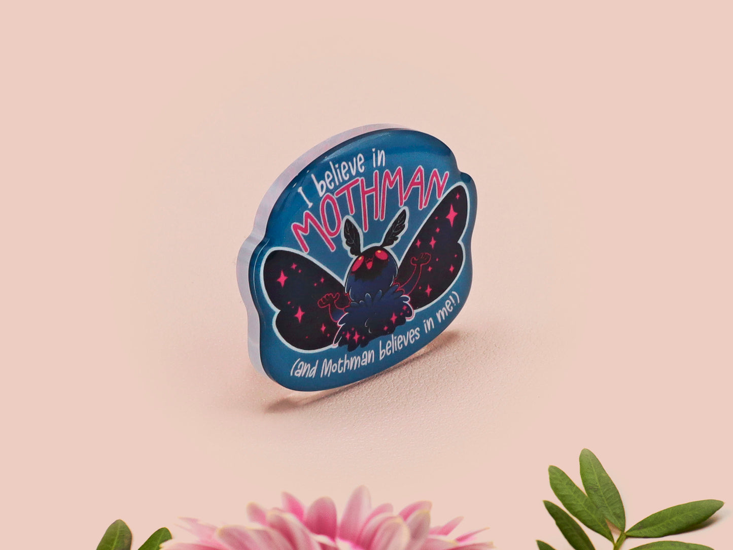 Clear acrylic pin badge with pink heart-shaped rubber pin back clasp. The design of the pin badge is a cute cartoon chibi Mothman cryptid with the quote I believe in Mothman and Mothman believes in me.