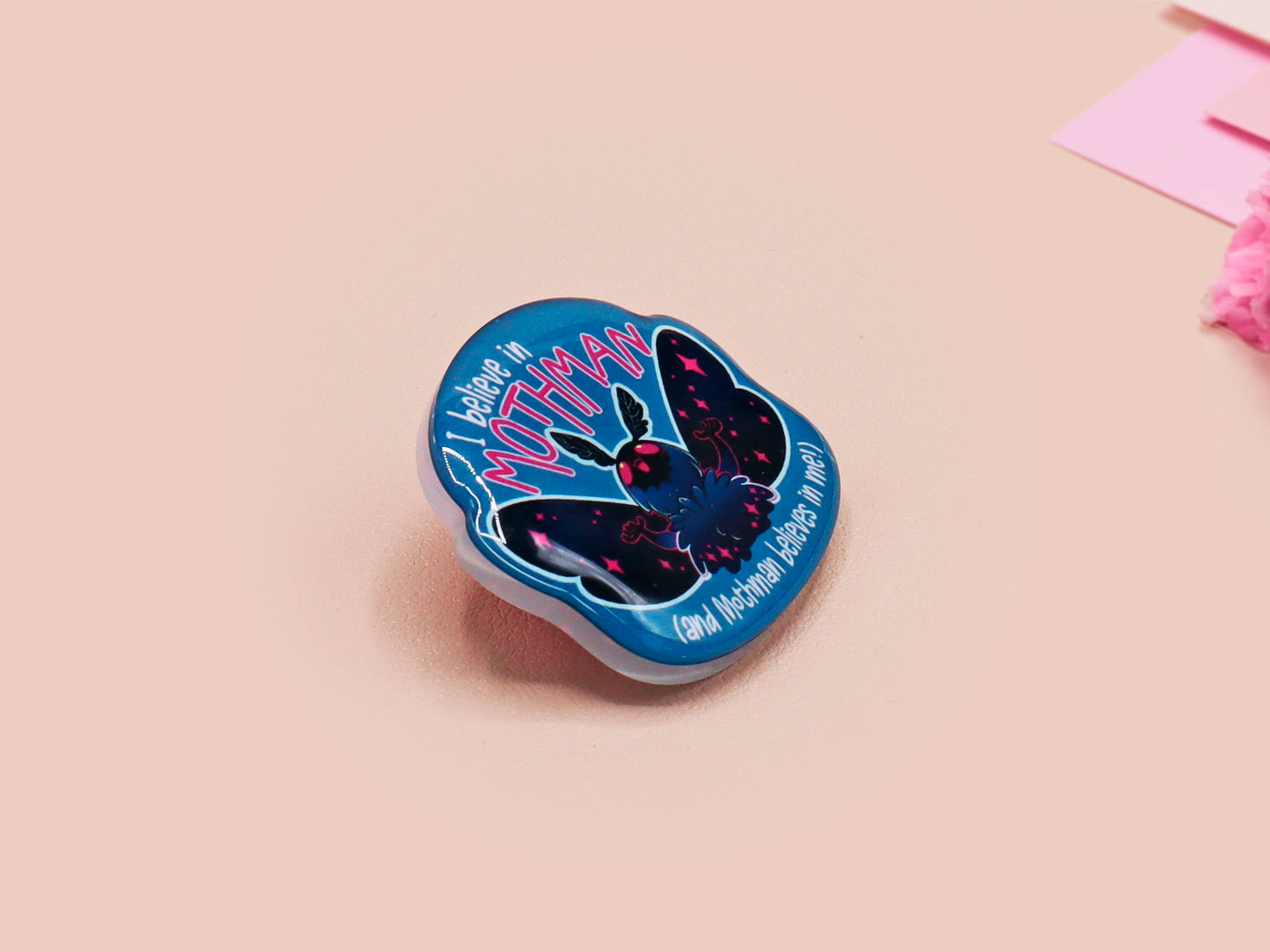 Clear acrylic pin badge with pink heart-shaped rubber pin back clasp. The design of the pin badge is a cute cartoon chibi Mothman cryptid with the quote I believe in Mothman and Mothman believes in me.