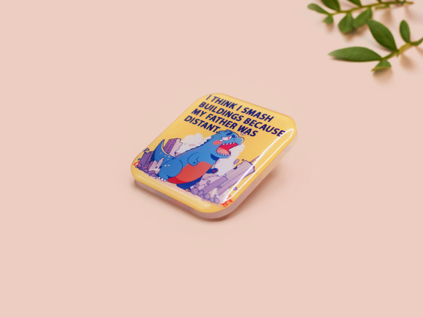 A clear acrylic pin badge with a pink heart-shaped pin back clasp. The design of the pin badge is of a cute chibi blue godzilla like kaiju destroying a city with the quote I Think I Smash Buildings Because My Father Was Distant.