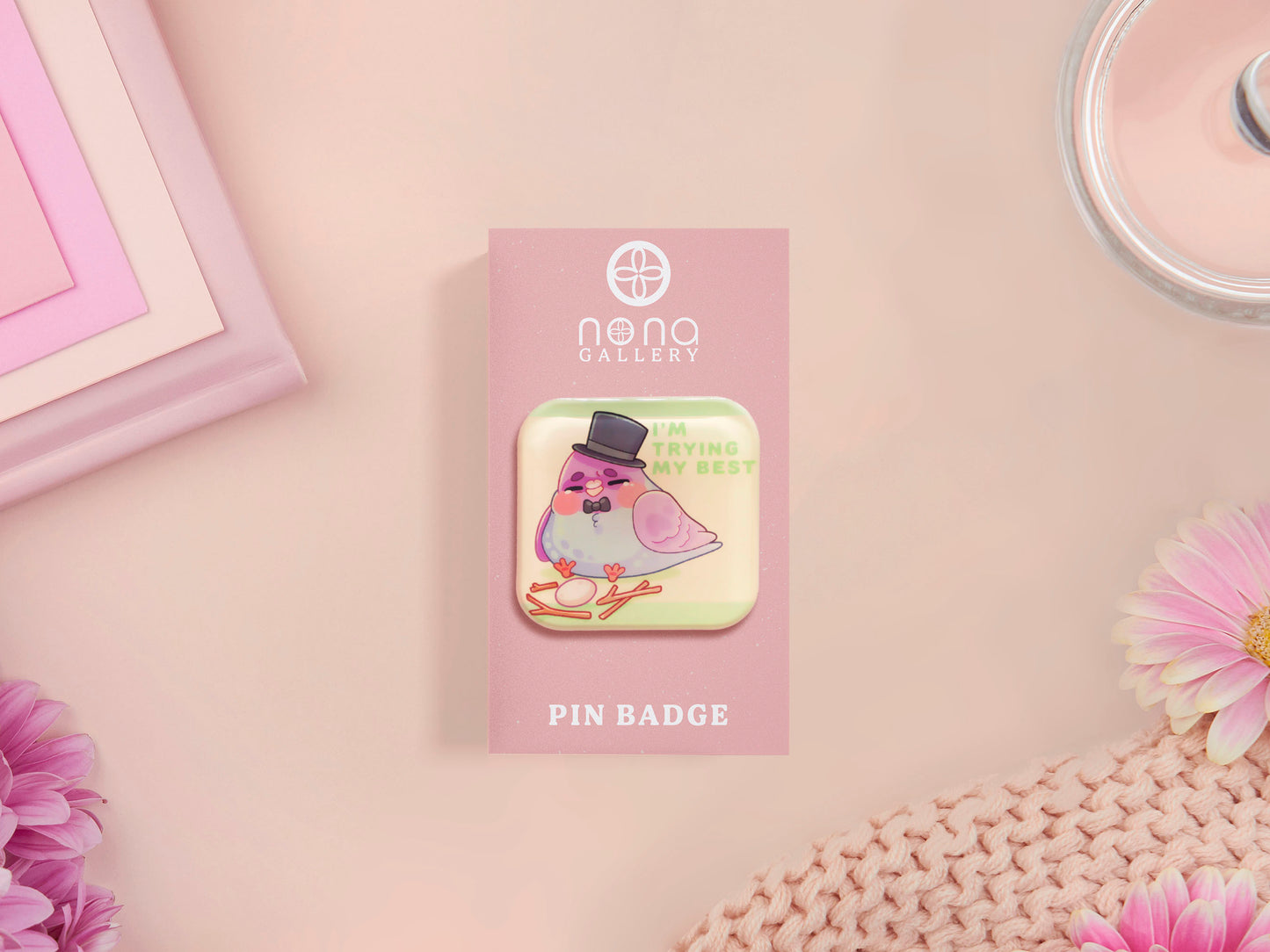 A clear acrylic pin badge with a pink heart-shaped pin back clasp. The design of the pin badge is of a cute chibi pigeon wearing a top hat crying next to a pethetic attempt at nest building with the quote I'm trying my best.