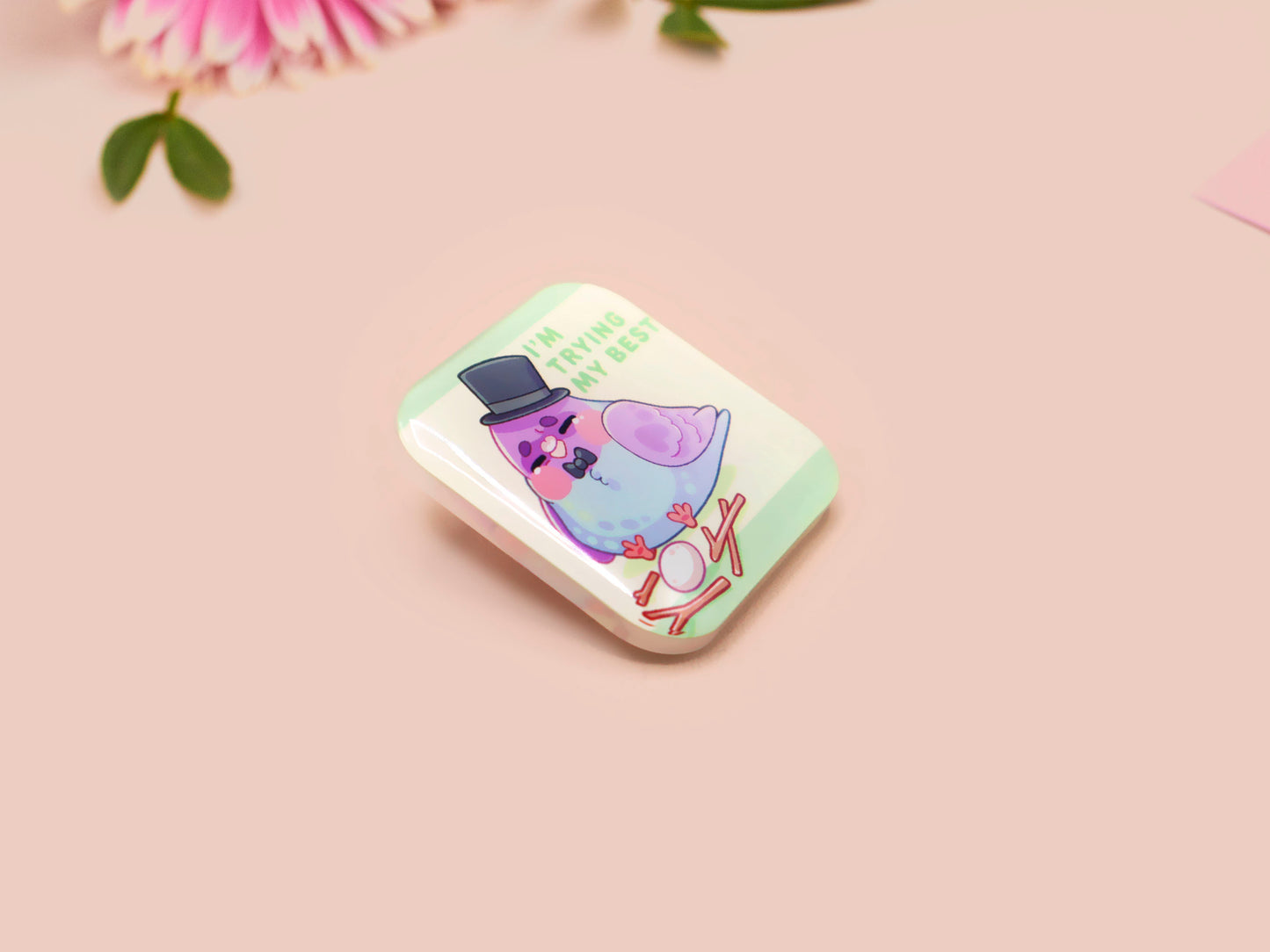 A clear acrylic pin badge with a pink heart-shaped pin back clasp. The design of the pin badge is of a cute chibi pigeon wearing a top hat crying next to a pethetic attempt at nest building with the quote I'm trying my best.