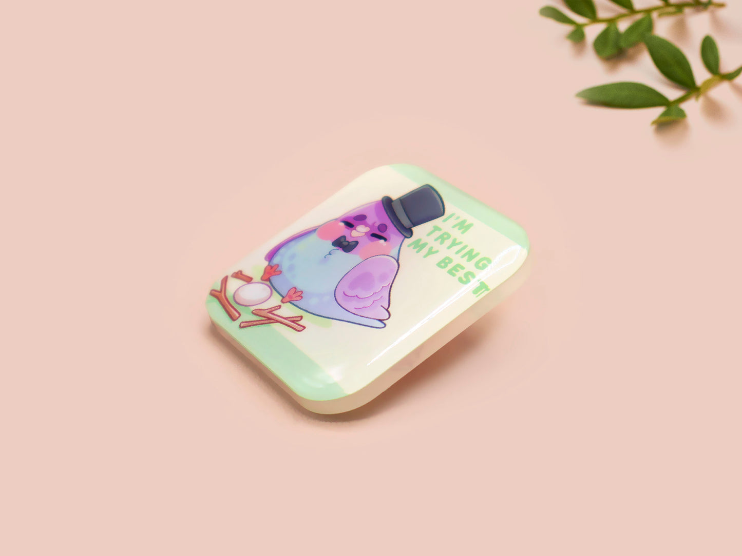 A clear acrylic pin badge with a pink heart-shaped pin back clasp. The design of the pin badge is of a cute chibi pigeon wearing a top hat crying next to a pethetic attempt at nest building with the quote I'm trying my best.