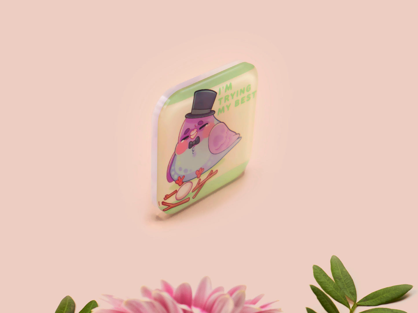 A clear acrylic pin badge with a pink heart-shaped pin back clasp. The design of the pin badge is of a cute chibi pigeon wearing a top hat crying next to a pethetic attempt at nest building with the quote I'm trying my best.