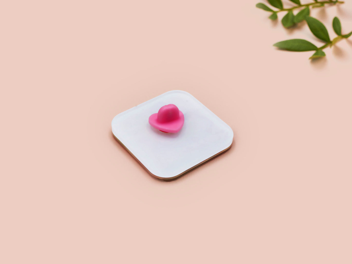 A clear acrylic pin badge with a pink heart-shaped pin back clasp. The design of the pin badge is of a cute chibi pigeon wearing a top hat crying next to a pethetic attempt at nest building with the quote I'm trying my best.