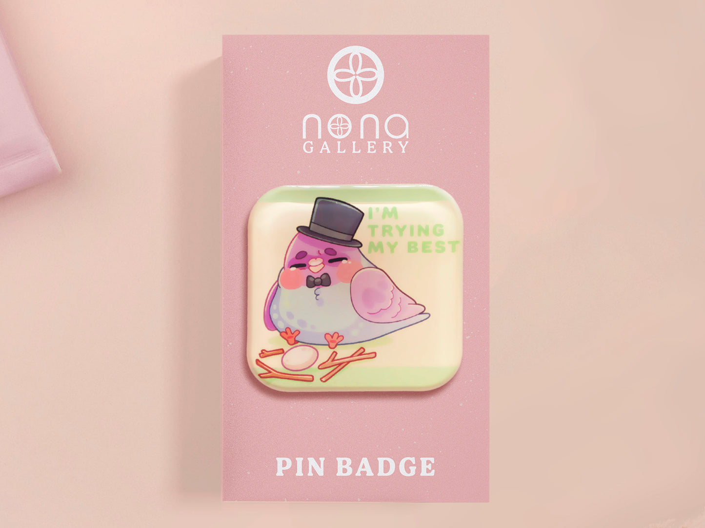 A clear acrylic pin badge with a pink heart-shaped pin back clasp. The design of the pin badge is of a cute chibi pigeon wearing a top hat crying next to a pethetic attempt at nest building with the quote I'm trying my best.