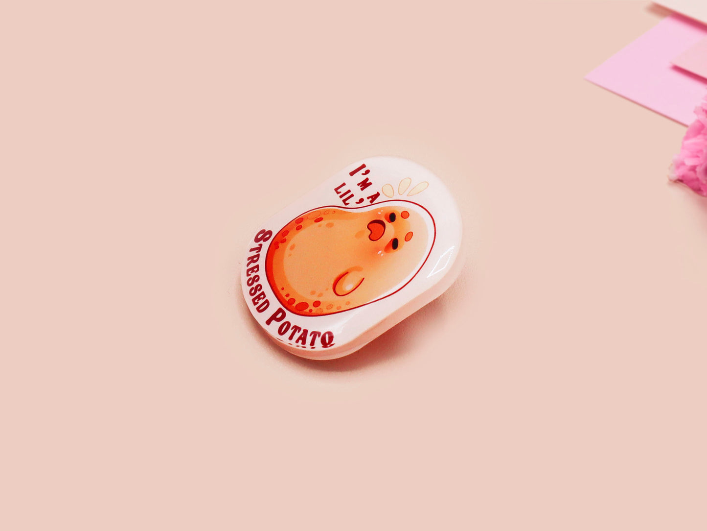 A clear acrylic pin badge with a pink heart-shaped pin back clasp. The design of the pin badge is of a cute chibi potato with the quote I'm a lil' stressed potato