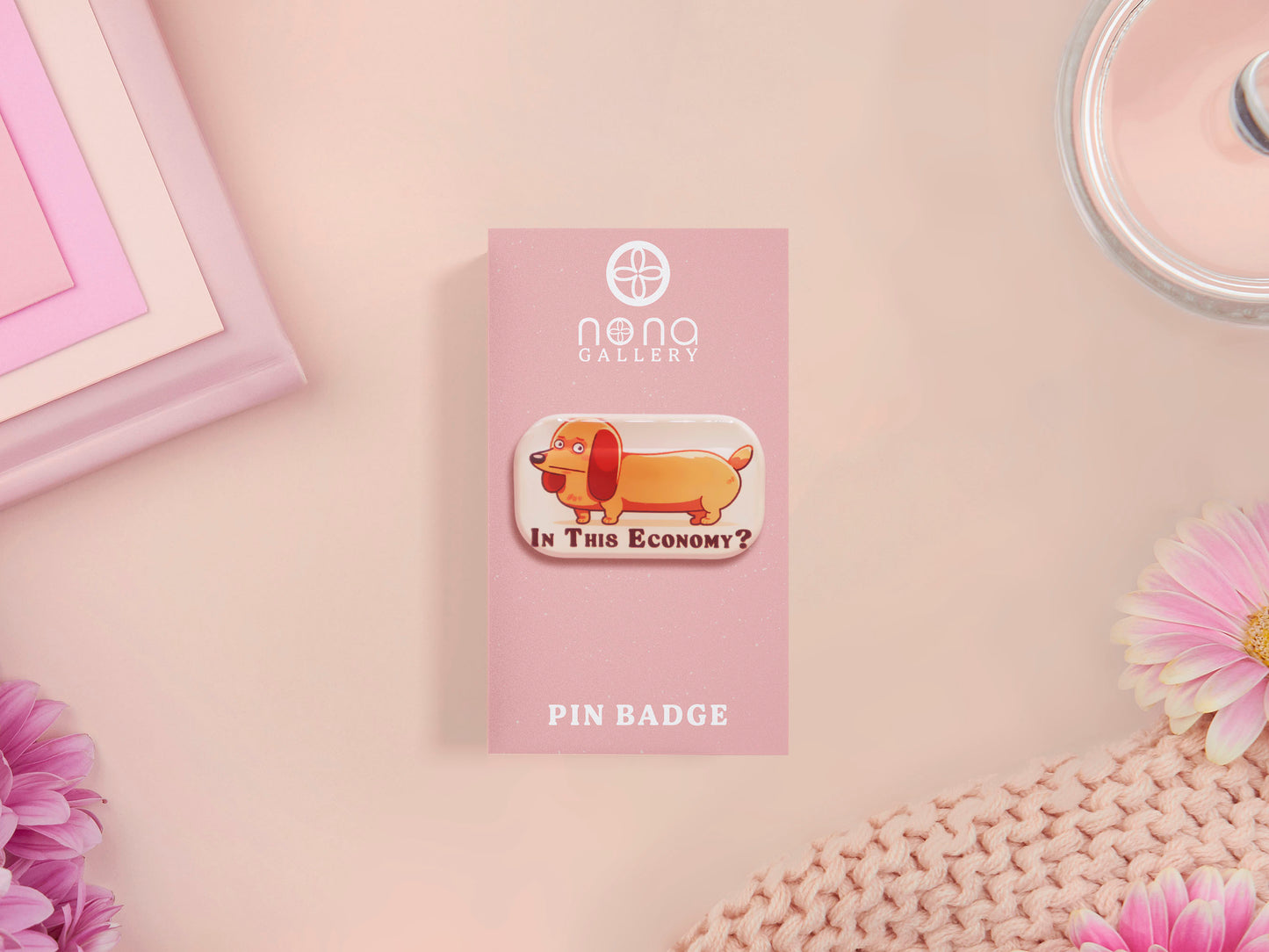 A clear acrylic pin badge with a pink heart-shaped pin back clasp. The design of the pin badge is of a cute chibi dachshund sausage dog with the quote in this economy?