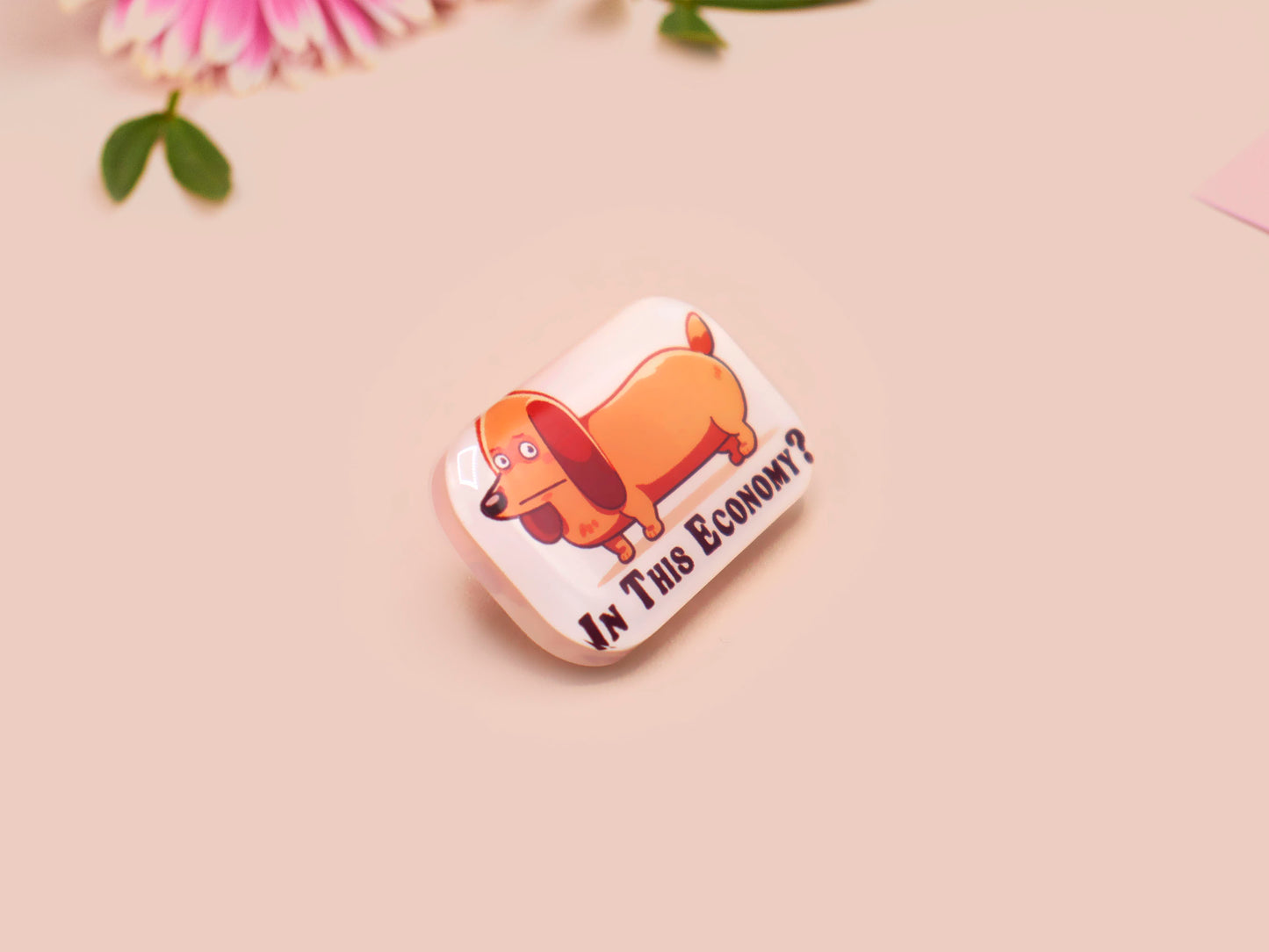 A clear acrylic pin badge with a pink heart-shaped pin back clasp. The design of the pin badge is of a cute chibi dachshund sausage dog with the quote in this economy?