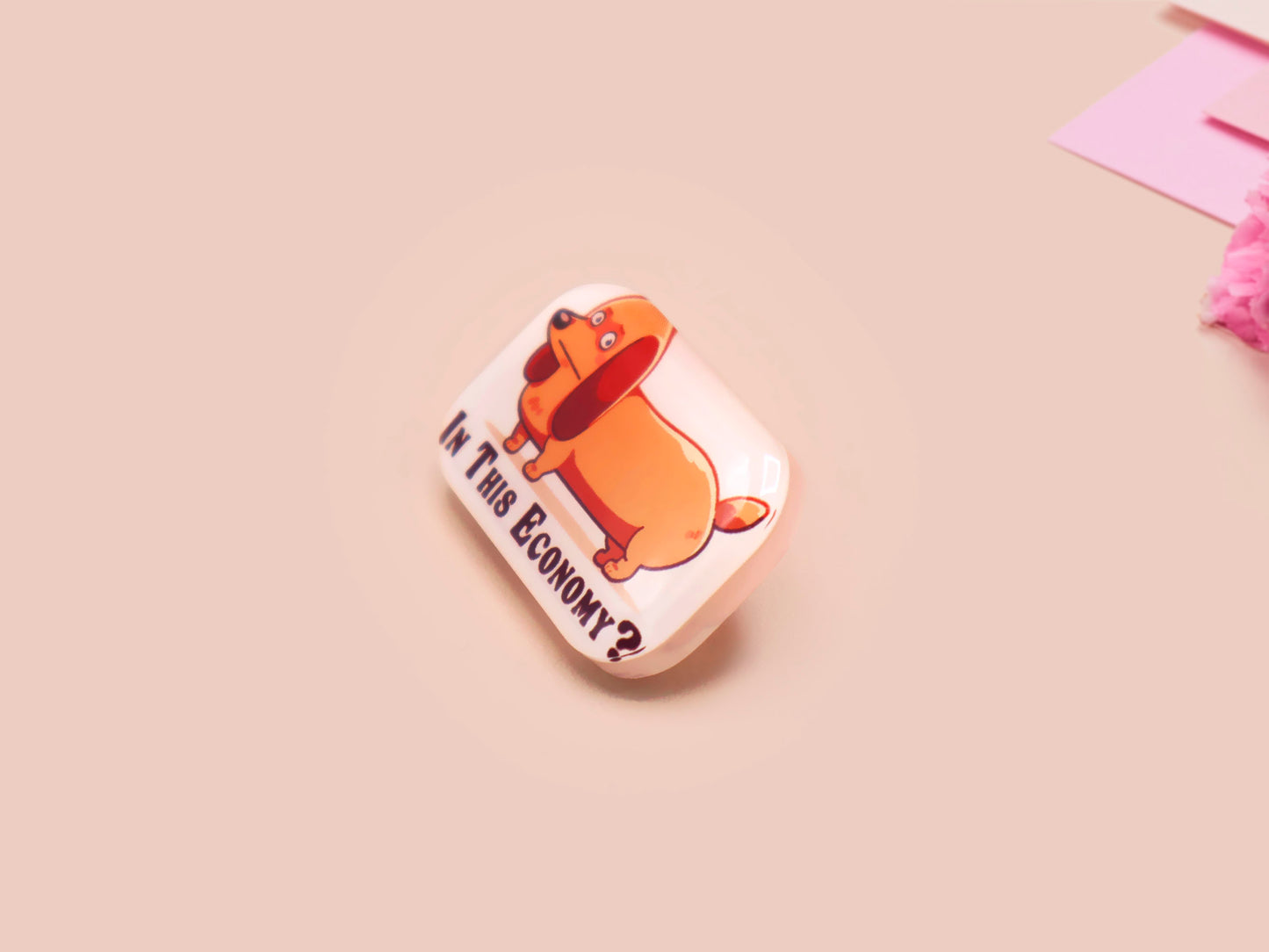 A clear acrylic pin badge with a pink heart-shaped pin back clasp. The design of the pin badge is of a cute chibi dachshund sausage dog with the quote in this economy?