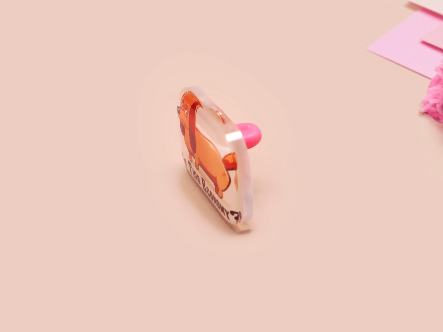 A clear acrylic pin badge with a pink heart-shaped pin back clasp. The design of the pin badge is of a cute chibi dachshund sausage dog with the quote in this economy?