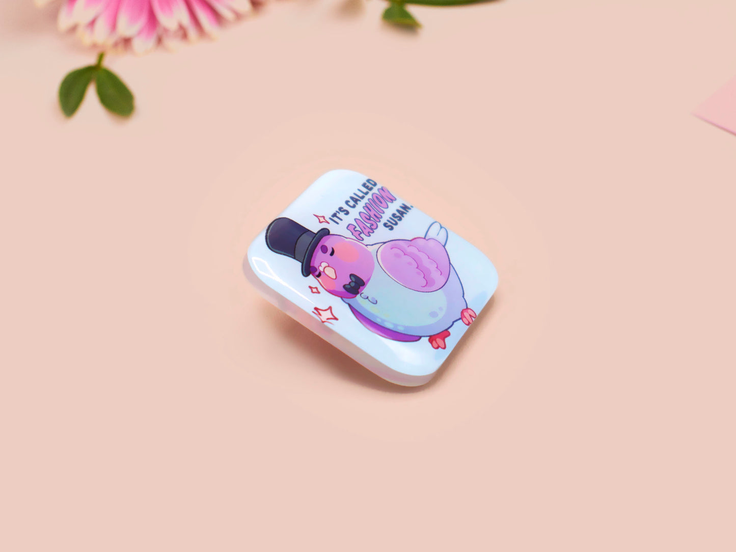 A clear acrylic pin badge with a pink heart-shaped pin back clasp. The design of the pin badge is of a cute chibi pigeon wearing a top hat with the quoteit's called fashion susan