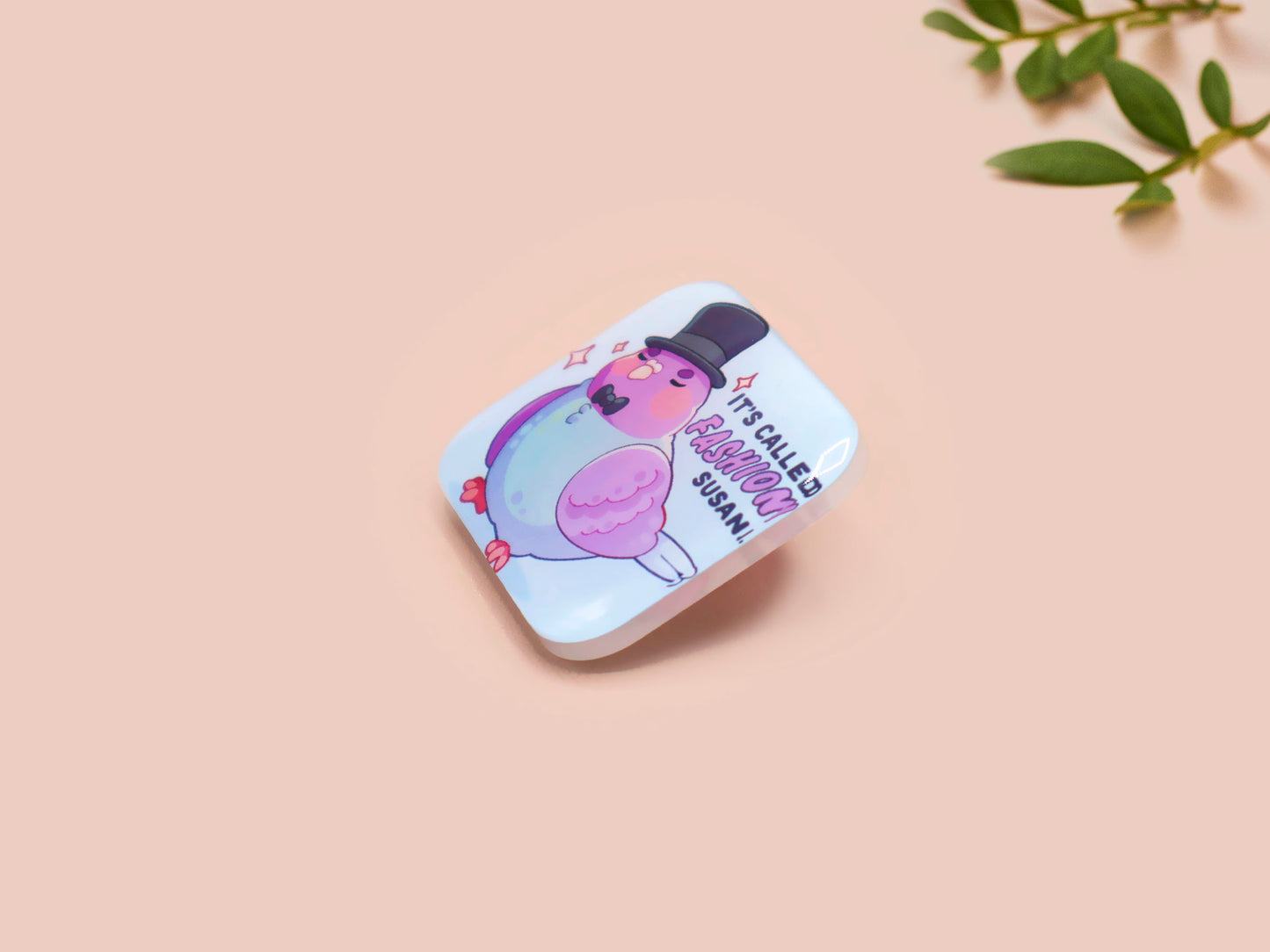 A clear acrylic pin badge with a pink heart-shaped pin back clasp. The design of the pin badge is of a cute chibi pigeon wearing a top hat with the quoteit's called fashion susan