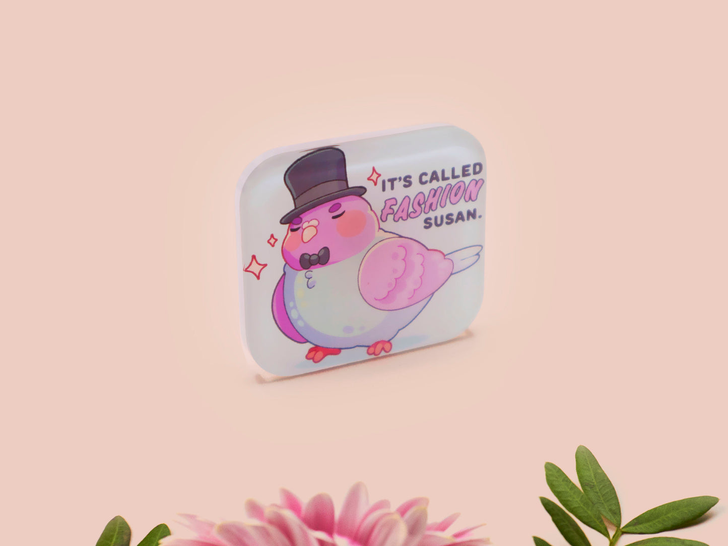 A clear acrylic pin badge with a pink heart-shaped pin back clasp. The design of the pin badge is of a cute chibi pigeon wearing a top hat with the quoteit's called fashion susan