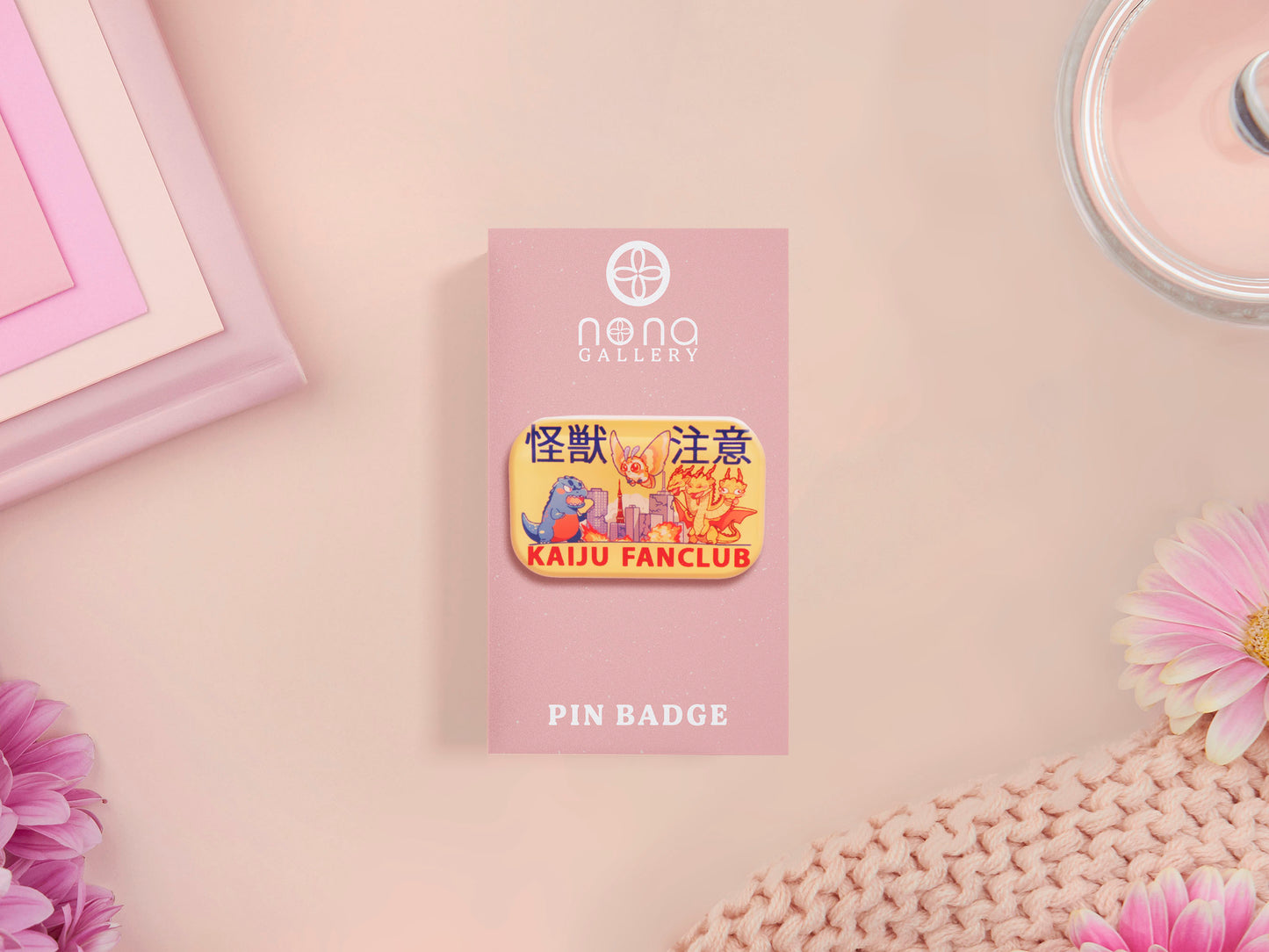 A clear acrylic pin badge with a pink heart-shaped pin back clasp. The design of the pin badge is of a cute chibi blue godzilla like kaiju mothra and King Ghidorah destroying a city with the quote kaiju fanclub and kaiju warning in japanese kanji