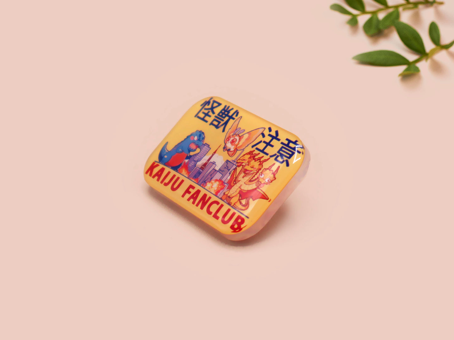 A clear acrylic pin badge with a pink heart-shaped pin back clasp. The design of the pin badge is of a cute chibi blue godzilla like kaiju mothra and King Ghidorah destroying a city with the quote kaiju fanclub and kaiju warning in japanese kanji